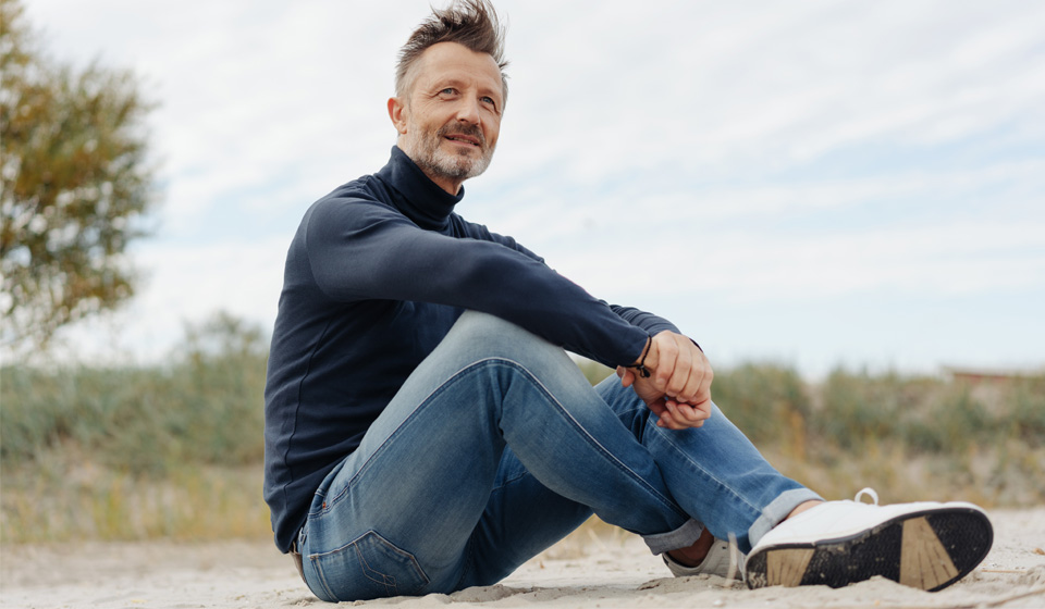 Een persoon zit buiten in een ontspannen houding op een zandige ondergrond, met een blauwe trui en jeans, tegen de achtergrond van een natuurlijke omgeving.