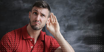Een man in een rood poloshirt houdt zijn hand achter zijn oor, alsof hij moeite heeft met horen. De achtergrond is een donkere krijtbord-achtige muur.