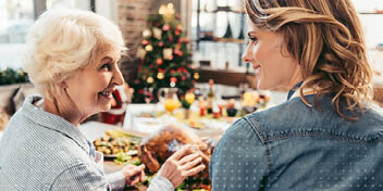 Een oudere vrouw en een jongere vrouw (gezichten geblurd) zitten samen aan een feestelijk gedekte tafel met eten, waaronder een kalkoen, in een warme huiselijke setting met een kerstboom op de achtergrond.