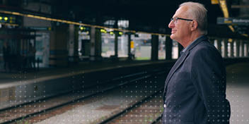 Een oudere man staat stil op een treinstation, kijkend naar de sporen. De omgeving toont perronborden, rails en lichtbalken.