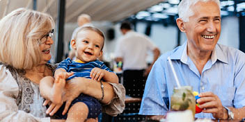 Een oudere man en vrouw zitten aan een tafeltje met een baby op schoot in een open restaurantomgeving. De man houdt een kleurrijke bal vast; achtergrond toont mensen en verlichting.