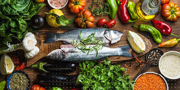 Twee hele vissen liggen op een houten plank, gegarneerd met kruiden, omringd door verse groenten (paprika's, tomaten, spinazie), citrusvruchten, specerijen, en kookingrediënten in een kleurrijke keukenopstelling.