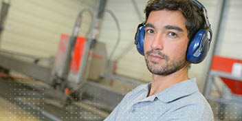 Een man draagt geluiddempende koptelefoon en bevindt zich in een industriële werkplaats met machines op de achtergrond.