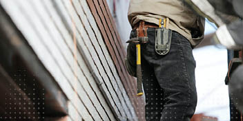 Een bouwvakker loopt op een schuin dak, met een hamer in een gereedschapshouder aan zijn broek. De omgeving toont bakstenen en metalen dakbedekking in daglicht.