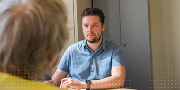 Een audioloog in een blauw overhemd zit aan een tafel en spreekt met een oudere persoon in een gele trui. De professionele omgeving suggereert een hoorkliniekconsult.