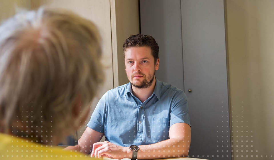 Een audioloog in een blauw overhemd zit tegenover een oudere persoon in een gele trui tijdens een consult in een hoorcentrum. De omgeving bestaat uit kasten en een rustige, professionele ruimte.