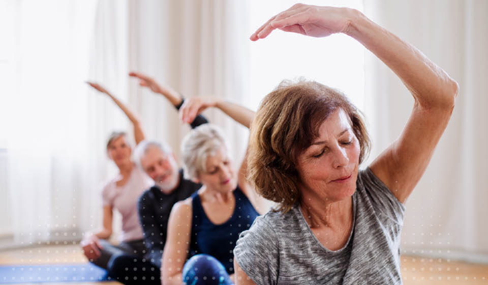 Een groep oudere vrouwen voert stretch- of yogaoefeningen uit, zittend op een mat in een lichte ruimte met grote ramen en gordijnen; gezichten zijn geblurd.