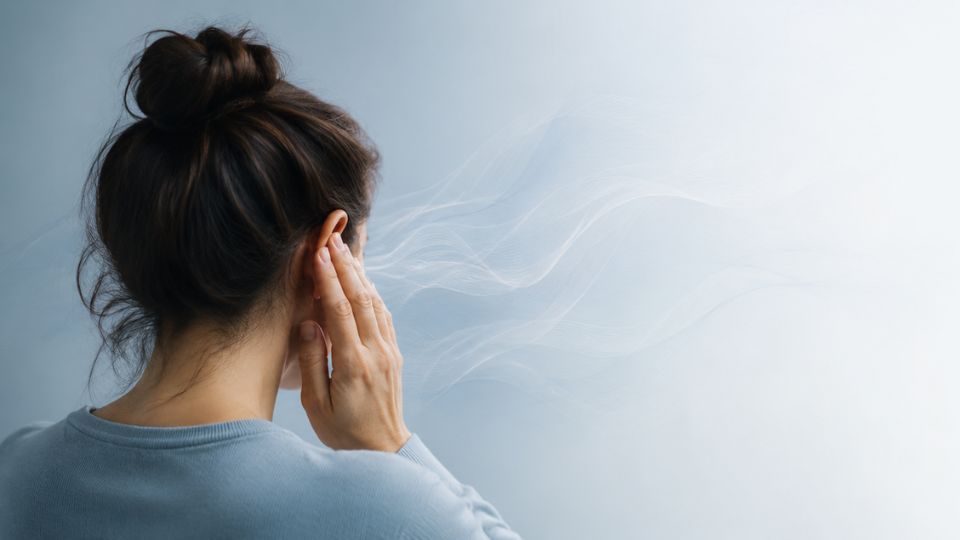 Een vrouw gezien van achteren houdt haar hand tegen haar oor, alsof ze gehoorproblemen of plotsdoofheid ervaart. Vanuit haar oor lijken zachte, golvende lijnen te komen die geluid symboliseren, tegen een lichte, minimalistische achtergrond.