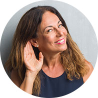 Een vrouw met halflang bruin haar legt haar hand achter haar oor, alsof ze luistert, tegen een neutrale grijze achtergrond.