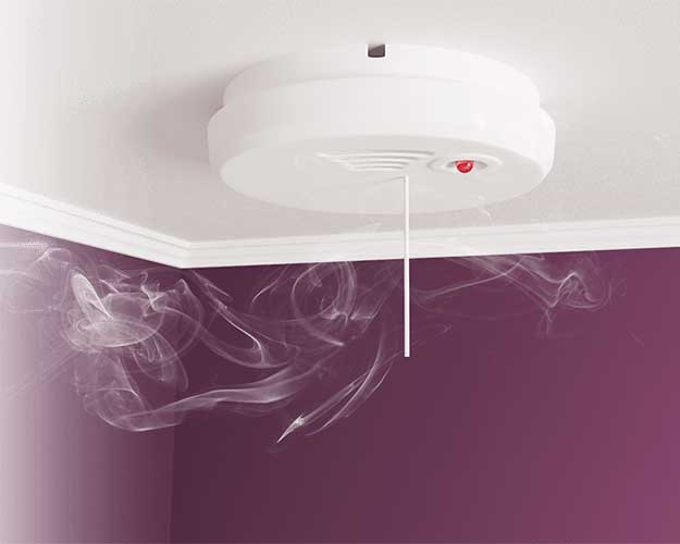 Een witte rookmelder aan een plafond detecteert rook, zichtbaar door rookwolken die opstijgen in een kamer met paarse muren en witte plafondlijsten. 