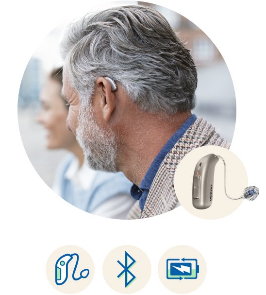 Oticon More-hoortoestel gedragen door een man met grijs haar, met symbolen voor connectiviteit, Bluetooth en oplaadbaarheid.
