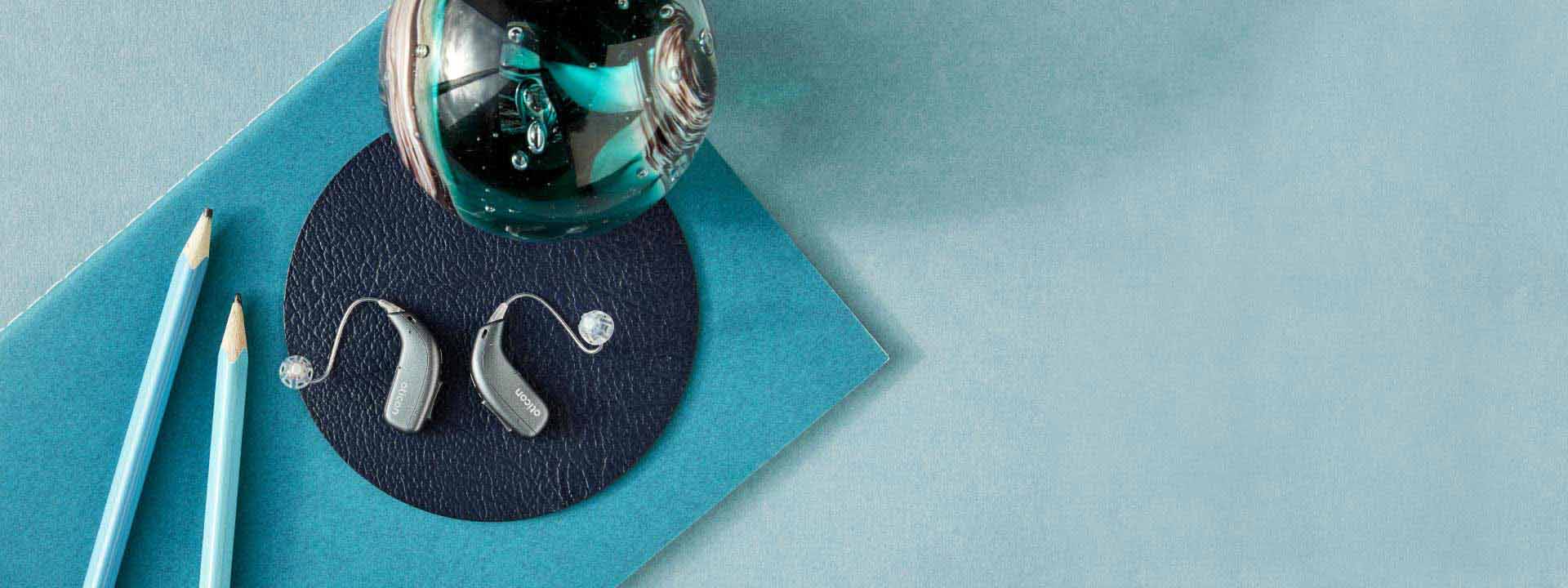 Oticon More hoortoestellen op een blauwe ondergrond met potloden en decoratief object.