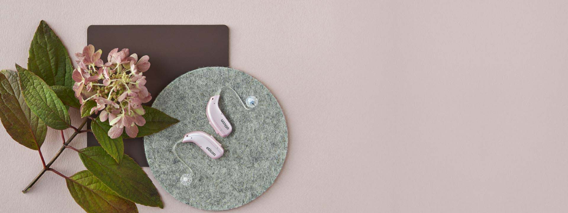 Twee roze Oticon More hoortoestellen op een ronde vilten ondergrond naast een hortensiabloem en groene bladeren.