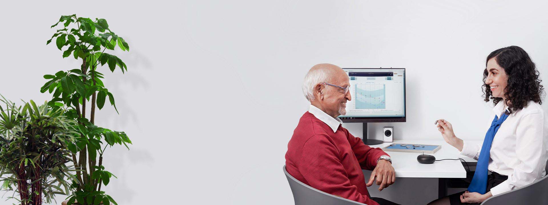 Audicien voert een gesprek met een oudere man met gehoorverlies terwijl een audiogram wordt weergegeven op een scherm.