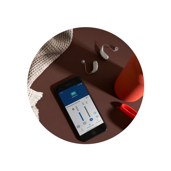 Hoorapparaten naast een smartphone met gehoorapparaatinstellingen op het scherm, liggend op een bruine ondergrond.