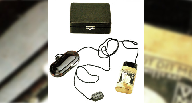 Een vintage hoortoestel met kabels, een batterijhouder met foto van een vrouw, en een zwart opbergdoosje. Ideaal voor historische contexten in gehoorapparaat-technologie.
