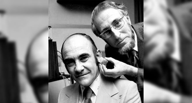 Een audioloog past een hoortoestel aan bij een zittende man in een professionele omgeving, mogelijk een hoorcentrum. De setting bevat apparatuur die hoortests ondersteunt. Geen merk zichtbaar.