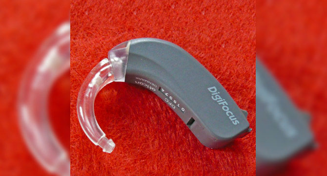 Een grijs Oticon Digifocus-hoortoestel met een transparante oorhaak ligt op een felrode, harige ondergrond. Het logo 'Digifocus' en technische details zijn op het apparaat zichtbaar.