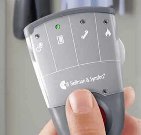 Een hand houdt een zilvergrijs apparaat vast met het logo 'Bellman & Symfon'. Het apparaat toont pictogrammen: telefoon, batterij, vlam en bel. Een groene LED is geactiveerd.