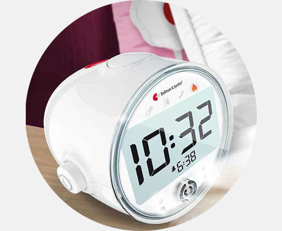 Een ronde, witte wekker van Bellman & Symfon toont de tijd '10:32' op een digitaal scherm. Het apparaat bevindt zich op een houten nachtkastje naast bedtextiel.