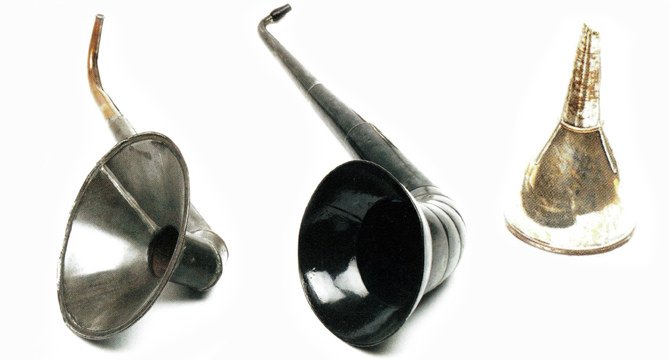 Drie oude akoestische hoortoestellen worden getoond: een metalen hoorn, een langwerpige zwarte hoorn en een kleine, taps toelopende hoorn. Ze liggen tegen een witte achtergrond.