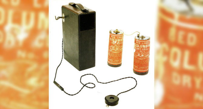 Een zwart, rechthoekig hoortoestel is verbonden via draden aan twee rode 'Red Label Columbia Dry Cell'-batterijen. De setting lijkt geïnspireerd door vroege gehoorhulpmiddelen in een historische context.