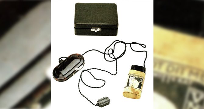 Een vintage hoortoestel met kabels, een batterijhouder met foto van een vrouw, en een zwart opbergdoosje. Ideaal voor historische contexten in gehoorapparaat-technologie.