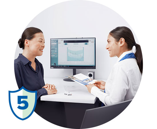 Een audioloog in witte jas bespreekt een hooradvies met een klant aan een bureau in een hoorcentrum. Op de monitor staat een audiogram. Linksonder staat een blauw schild met het getal 5.