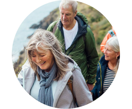 Eine Gruppe älterer Menschen wandert entlang einer Küstenlandschaft mit grünem Gelände und Meer im Hintergrund. Die Gesichter sind verpixelt, keine Hörgeräte oder Markenlogos sind erkennbar.