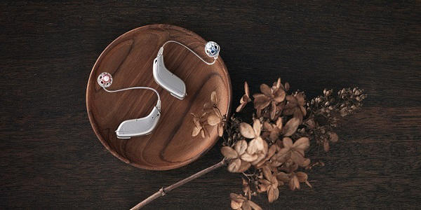 Nowoczesne aparaty słuchowe Oticon Opn™ na drewnianej misce z suszonymi kwiatami w tle.