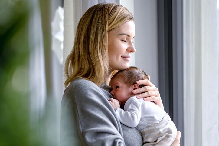 Kobieta trzymająca niemowlę przy oknie w jasnym pomieszczeniu.