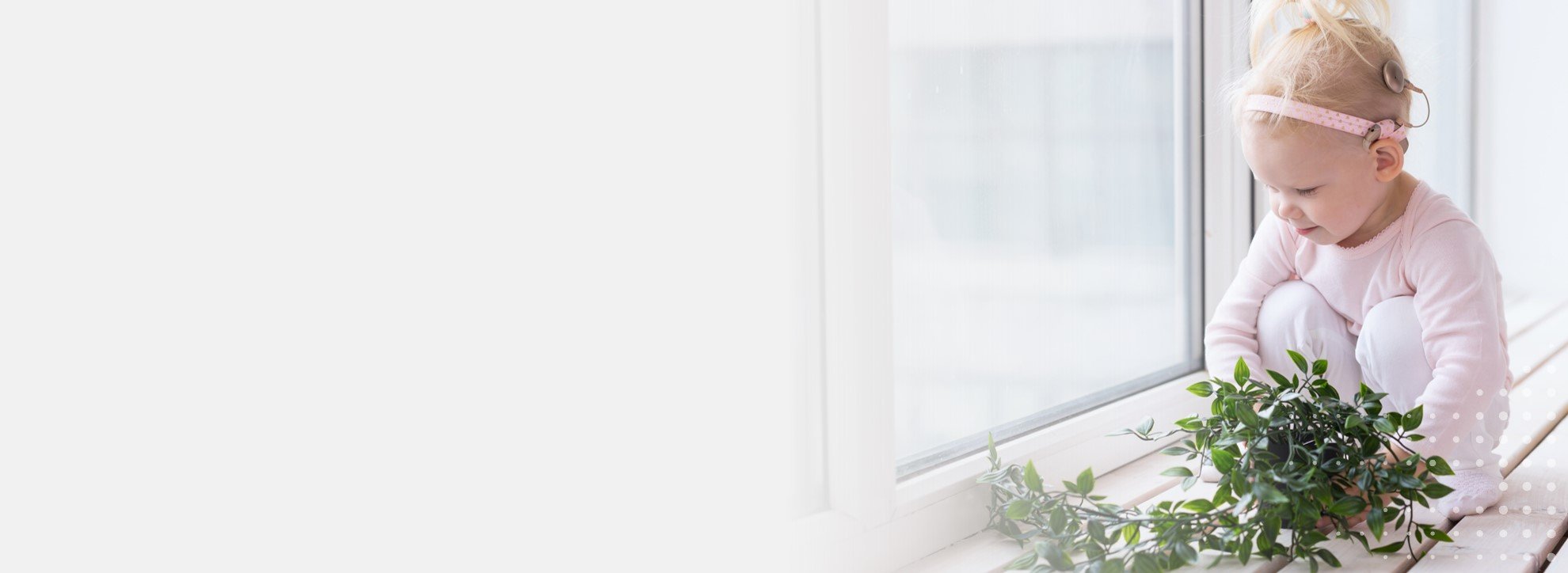 Dziewczynka z implantem słuchowym w różowej opasce siedzi na parapecie obok zielonej rośliny.