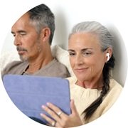 Starsza kobieta z szarym warkoczem korzystająca z aparatu słuchowego, siedząca obok mężczyzny, trzymającego tablet.