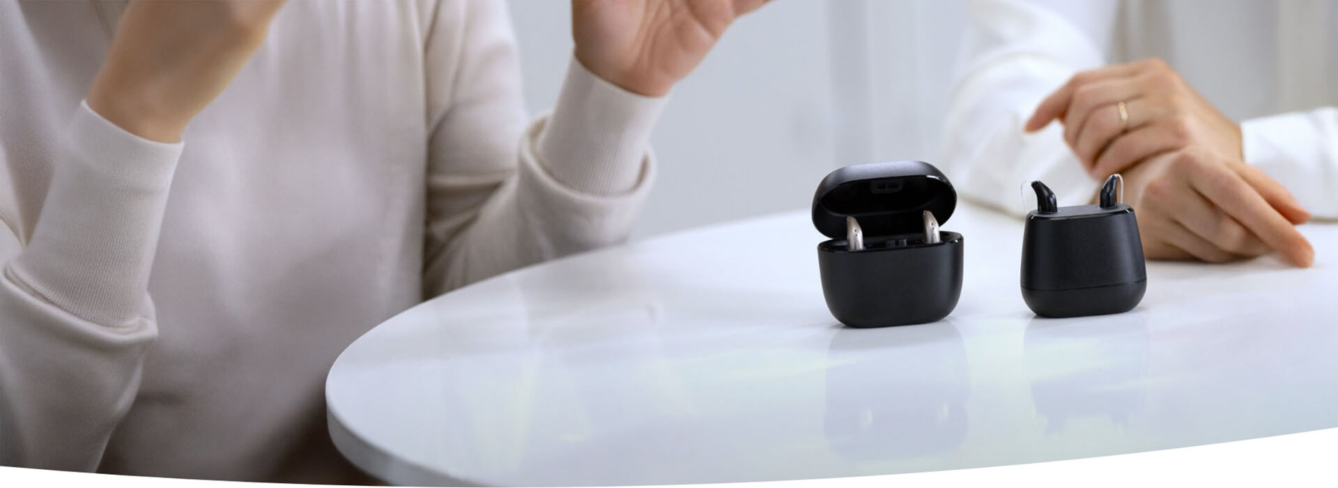 Nowoczesne aparaty słuchowe w etui na stole, w tle rozmowa osób.