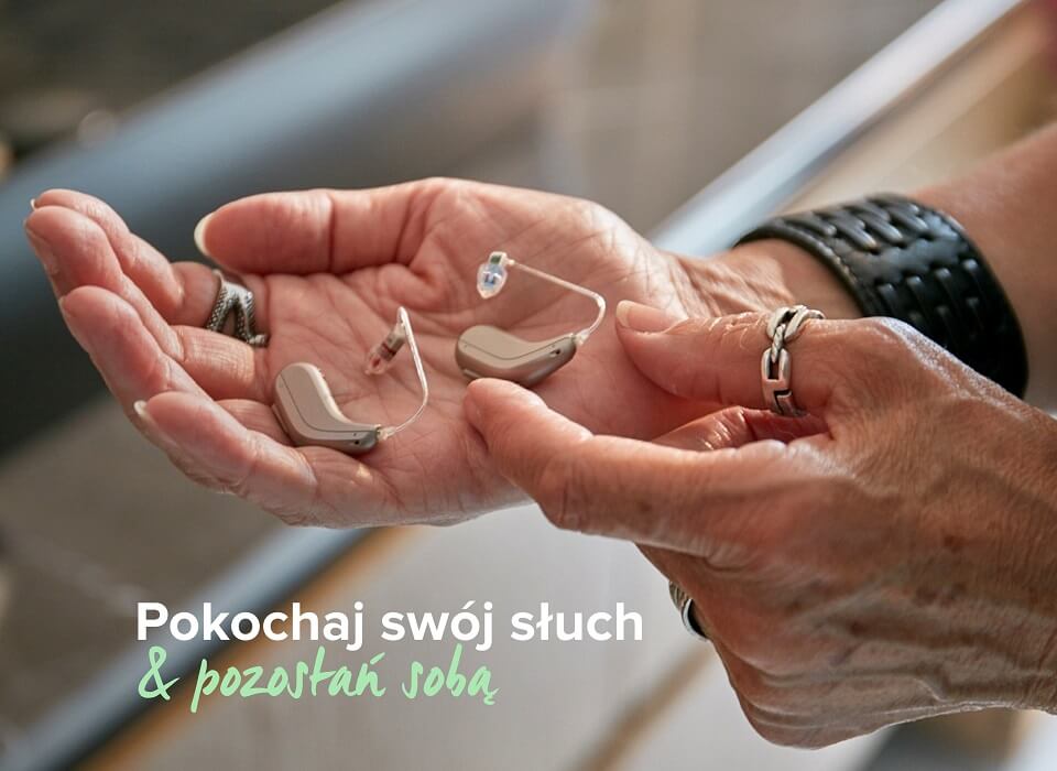 Dłonie trzymają dwa aparaty słuchowe, z widoczną delikatną konstrukcją. W tle zamazana przestrzeń. Tekst: „Pokochaj swój słuch & pozostań sobą”. Aparaty sugerują troskę o słuch.