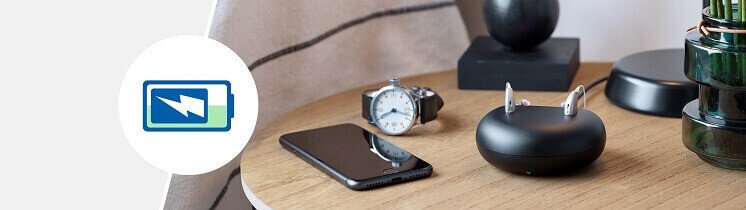 Aparat słuchowy Oticon Zircon™ z ładowarką na stoliku obok telefonu, zegarka i dekoracji.