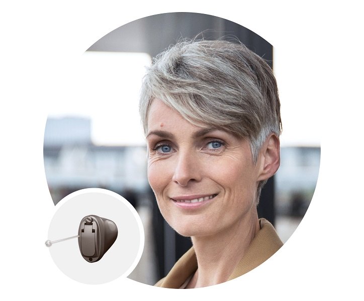 Kobieta z krótkimi siwymi włosami nosi douszny aparat słuchowy ITE, stojąc na zewnątrz.