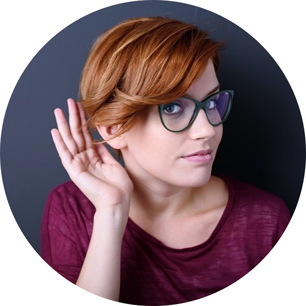 Młoda kobieta w okularach i bordowej bluzce wykonuje gest nasłuchiwania, dotykając dłonią ucha.