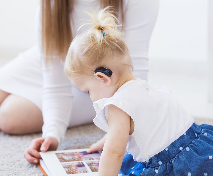 Dziecko noszące aparaty słuchowe przegląda książkę z mamą w domu.
