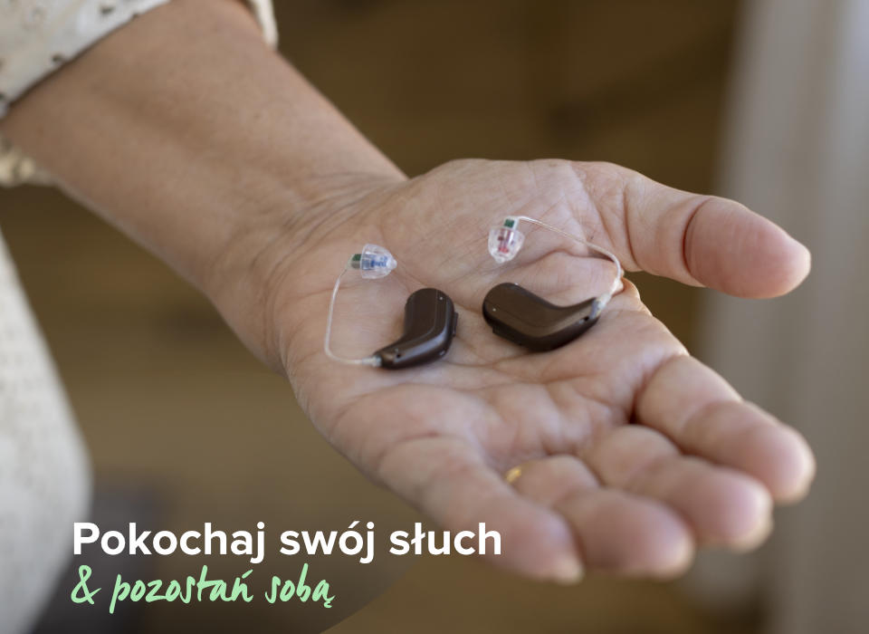 Dłonie trzymają dwa aparaty słuchowe, z widoczną delikatną konstrukcją. W tle zamazana przestrzeń. Tekst: „Pokochaj swój słuch & pozostań sobą”. Aparaty sugerują troskę o słuch.