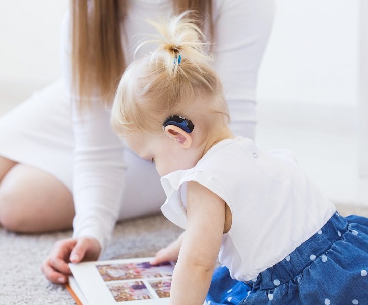 Dziecko noszące aparaty słuchowe przegląda książkę z mamą w domu.