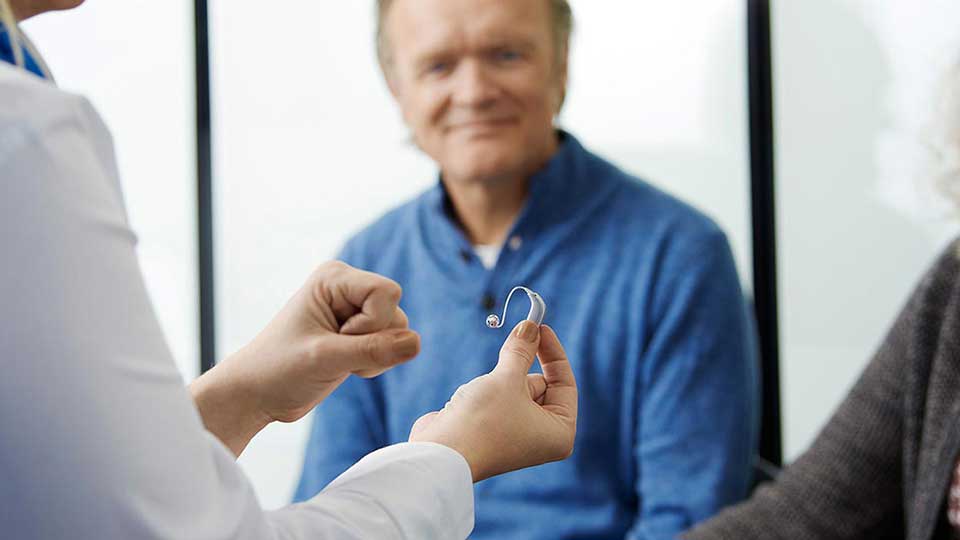  Kvinna som håller upp en hörapparat och berättar om fördelarna med att använda hörapparater och hur de kan förbättra din hörsel i vardagen. 