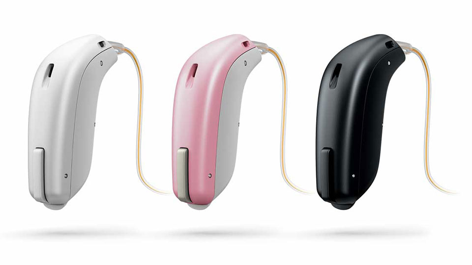  Tre hörapparater i olika färger – vit, rosa och svart – placerade på rad med tunna ljudslangar.