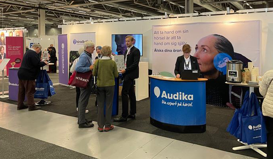  Audikas monter på en seniormässa med personal som pratar med besökare och skyltar som uppmuntrar till att ta hand om sin hörsel.