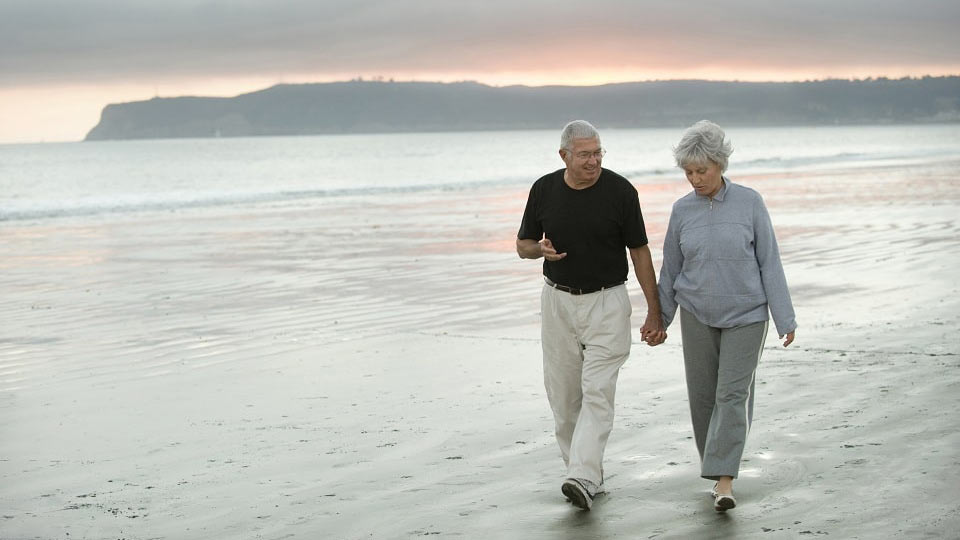  Bild på ett par som går på en strand och skyddar sina hörapparater mot fukt och värme under sommaren – 5 tips från Audika för att vårda dina hörapparater.