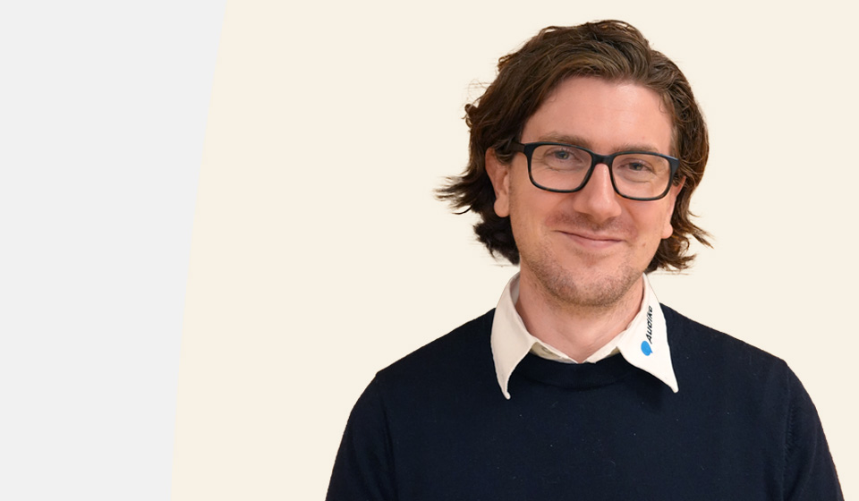  En audionom klädd i en mörk tröja över en vit skjorta med Audika-logotyp tryckt på kragen står mot en ljus beige bakgrund.