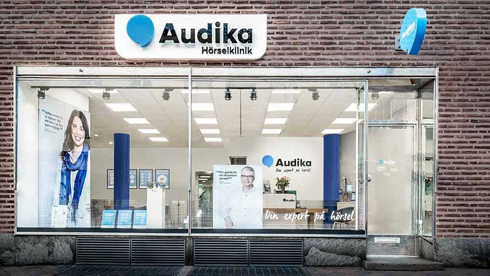  Audikas hörselklinik i Norrtälje med ingång och skyltfönster som visar klinikens interiör och slogan ’Din expert på hörsel’.