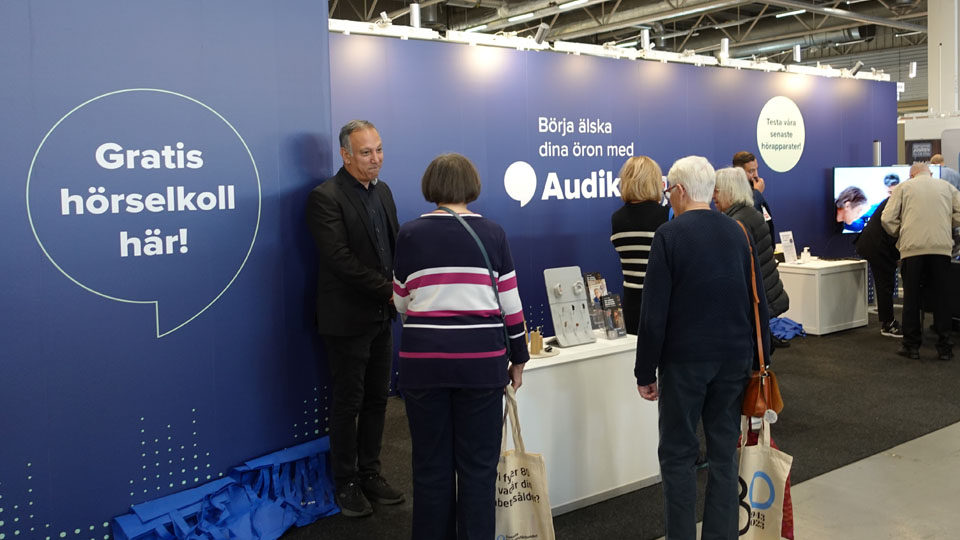  Audikas legitimerade audionomer pratar med besökare vid en monter på ett seniorevent med erbjudande om gratis hörselkoll.