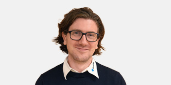  Person i mörk tröja med vit krage står mot en ljus bakgrund. En blå pin på kragen som är formad Audikas pratbubbla.