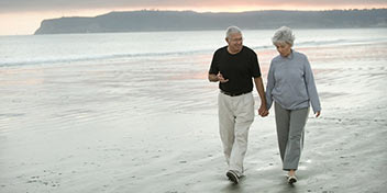  Äldre par promenerar hand i hand längs en strand i solnedgången.
